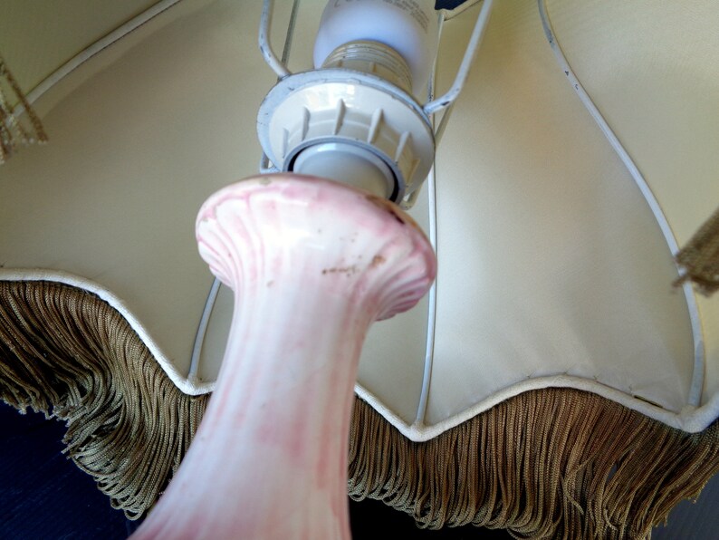 Op de afbeelding: Een roze keramische lampenvoet met een witte lampenkap van stof met een gouden franje. De lampenkap is bevestigd aan een metalen frame.