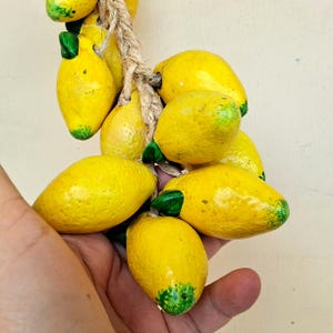 May include: A bunch of yellow ceramic lemons with green leaves hanging from a rope. The lemons are arranged in a cluster and are held in a person's hand.