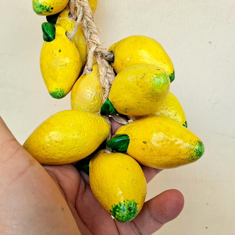 May include: A bunch of yellow ceramic lemons with green leaves hanging from a rope. The lemons are arranged in a cluster and are held in a person's hand.