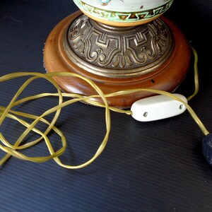 Op de afbeelding: Een vintage tafellamp met een keramische basis met een bloemenmotief. De lamp heeft een houten basis met een sierlijke metalen rand en een wit snoer met een schakelaar.