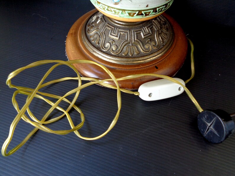 Op de afbeelding: Een vintage tafellamp met een keramische basis met een bloemenmotief. De lamp heeft een houten basis met een sierlijke metalen rand en een wit snoer met een schakelaar.