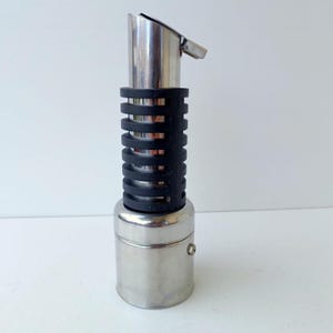 Puede incluir: Un vertedor de aceite o vinagre de acero inoxidable con una base cilíndrica y un cuello alto y estrecho. El cuello está envuelto en una empuñadura de silicona negra acanalada y la parte superior tiene un pico de metal inclinado y una tapa.