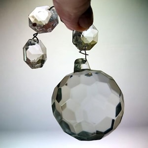 Può includere: Un ciondolo a sfera in vetro trasparente sfaccettato, collegato a due perline ottagonali in vetro più piccole tramite connettori in filo metallico scuro. Gli elementi in vetro presentano un taglio geometrico a più facce che riflette la luce.