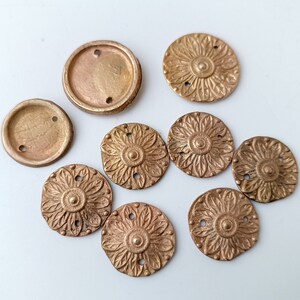 May include: A collection of antique bronze-colored metal embellishments. The assortment includes two circular pieces with two holes each, and several decorative floral-shaped pieces with intricate petal designs and small holes for attachment. The pieces are arranged on a white surface.