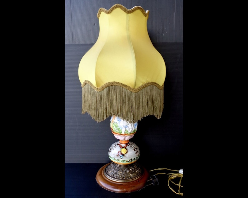 Op de afbeelding: Een vintage tafellamp met een gele lampenkap met gouden franjes. De basis is gemaakt van keramiek en heeft een kleurrijk bloemenmotief. De lamp heeft een houten voet en een gouden metalen accent.