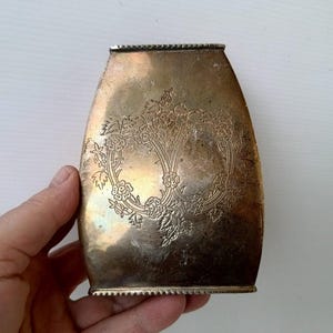 Puede incluir: Un recipiente de latón antiguo y deslustrado con un diseño floral. El recipiente tiene forma rectangular con bordes redondeados y un borde decorativo. El diseño floral está centrado en la parte delantera.