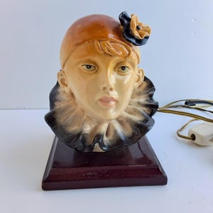May include: A vintage ceramic lamp shaped like a woman's bust. The lamp features a brown hat with a black and yellow flower, a ruffled collar, and a dark wooden base. The face is painted with neutral tones.