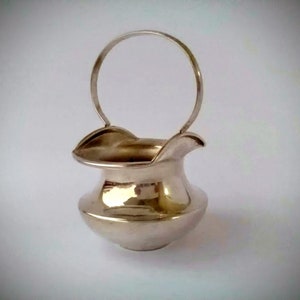 May include: A silver sugar bowl with a handle. The bowl has a rounded shape and a smooth, shiny surface.