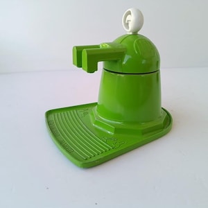 Puede incluir: Una cafetera espresso de color verde brillante con un pomo blanco en la parte superior. La base presenta una zona de bandeja de goteo estriada con el texto en relieve ESPRESSO MAKER. La unidad tiene un cuerpo cilíndrico central con un pico.