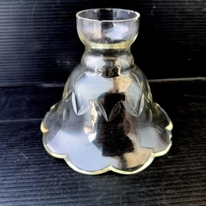 May include: A clear glass candle holder with a flared base and a scalloped edge. The body of the holder features etched leaf designs. The top has a narrow opening for a candle. The holder is sitting on a dark surface.