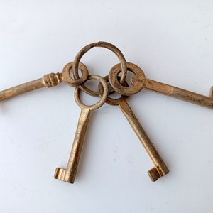 Op de afbeelding: Een set antieke messing sleutels aan een ring. De sleutels hebben een versleten, verouderde uitstraling, met zichtbare gebruikssporen. Ze zijn geplaatst tegen een witte achtergrond, waardoor hun details en vintage charme worden benadrukt.