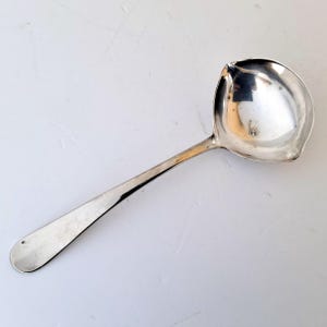 May include: A silver ladle with a long handle and a rounded bowl. The bowl has a pouring spout on one side. The ladle is made of a shiny, reflective metal, and it is set against a white background.