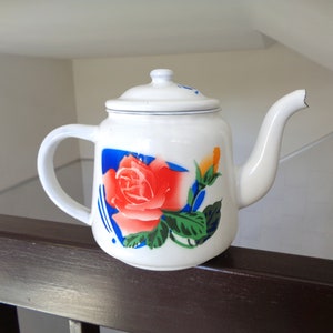 Könnte beinhalten: Weiße Keramik-Teekanne mit Deckel, Henkel und Ausguss. Die Teekanne ist mit einer großen roten Rose, grünen Blättern und blauen Akzenten verziert. Die Teekanne steht auf einer braunen Oberfläche.