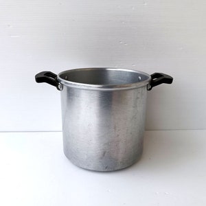 Puede incluir: Una olla grande y cilíndrica de aluminio con dos asas negras. La olla tiene un acabado de metal cepillado y está diseñada para cocinar. Las asas están unidas a los lados de la olla, proporcionando un agarre seguro. La olla está vacía y lista para usar.