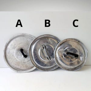 Puede incluir: Tres tapas de olla de metal vintage de diferentes tamaños. Las tapas son circulares con un acabado plateado y asas negras. Las tapas están etiquetadas A, B y C. Las tapas están dispuestas sobre una superficie blanca.