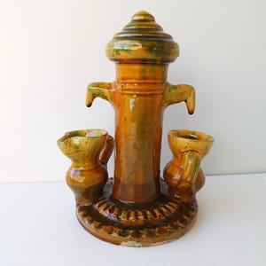 May include: A ceramic water fountain with a green and brown glaze. The fountain features a central column with a spout and two smaller jugs on either side.