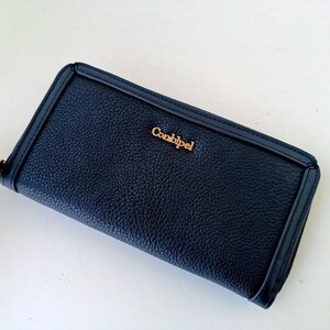 May include: A navy blue wallet with a textured surface and a gold-colored "Combipel" logo. The rectangular wallet has a zipper closure and a wrist strap. The wallet is a dark blue color.