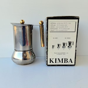 May include: A stainless steel stovetop espresso maker with a gold handle and accents. The box next to it has the brand name "KIMBA" and diagrams of different sizes. The text on the box lists the product in multiple languages.