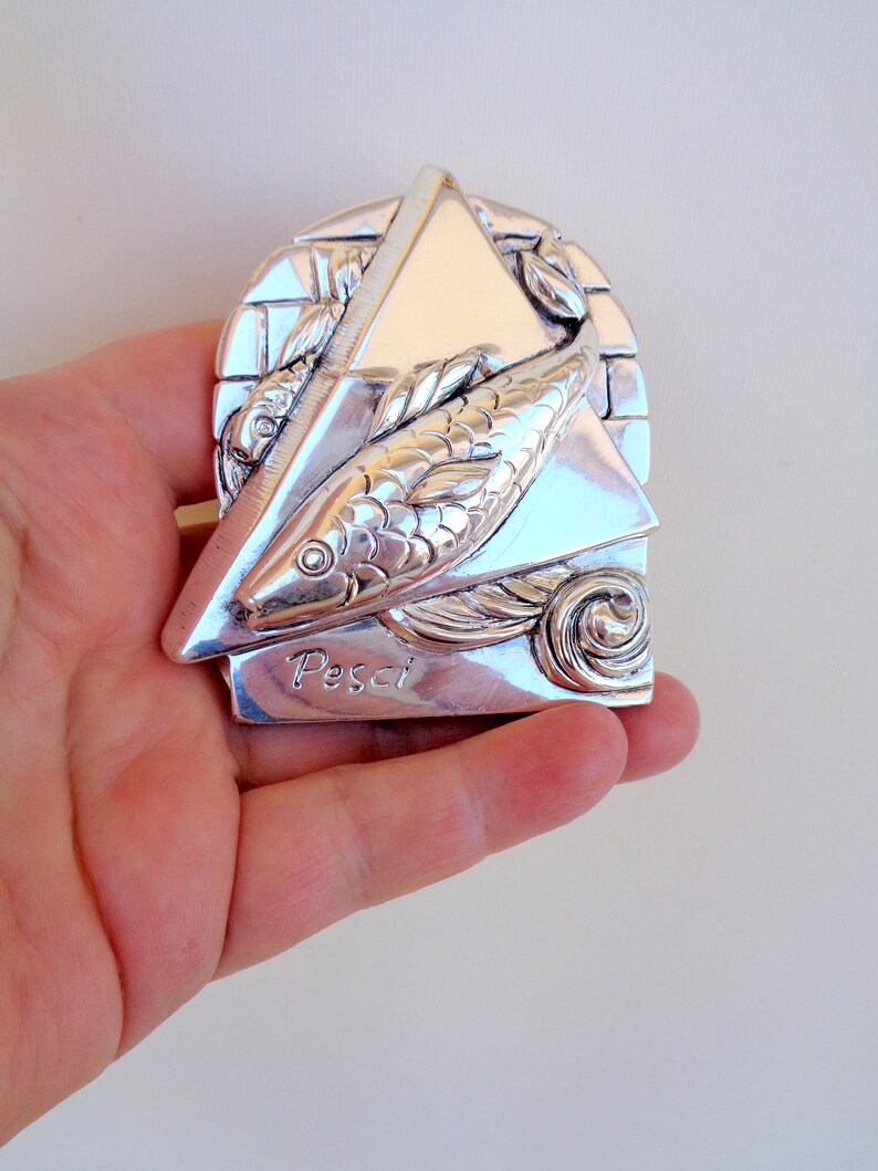 May include: Silver zodiac pendant featuring a stylised fish design with the word "Pisces" inscribed below. The pendant is in the shape of a triangle with a textured background.