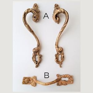 May include: Two antique brass drawer pulls and a matching handle. The drawer pulls are curved with ornate detailing and keyhole cutouts. The handle is a smaller, rectangular piece with a curved bar and floral accents. All pieces have a vintage, aged appearance.