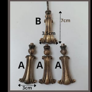 Può includere: Una vista dall'alto mostra quattro pomelli per mobili antichi color bronzo. Tre pomelli, etichettati "A", sono larghi 3 cm. Il quarto pomello, etichettato "B", è largo 3,5 cm e alto 7 cm. Tutti i pomelli presentano disegni decorativi e ornati.