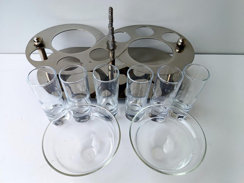 May include: A silver-toned serving set with a central handle and two oval trays with circular cutouts. It includes six clear shot glasses and two clear glass dishes, perfect for serving appetizers or drinks.