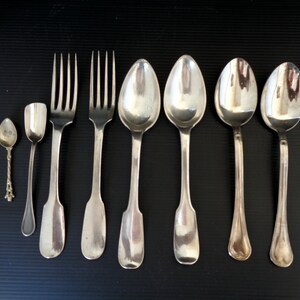 May include: A set of seven silver-plated utensils, including two forks, four spoons, and a small spoon. The utensils are arranged in a row on a black surface.