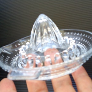 Op de afbeelding: Een heldere glazen citruspers met een geribbelde ruimer en een komvormige basis. De basis heeft een schenktuit en kleine, verhoogde bobbels rond de rand. De pers is ontworpen voor het handmatig persen van citrusvruchten.