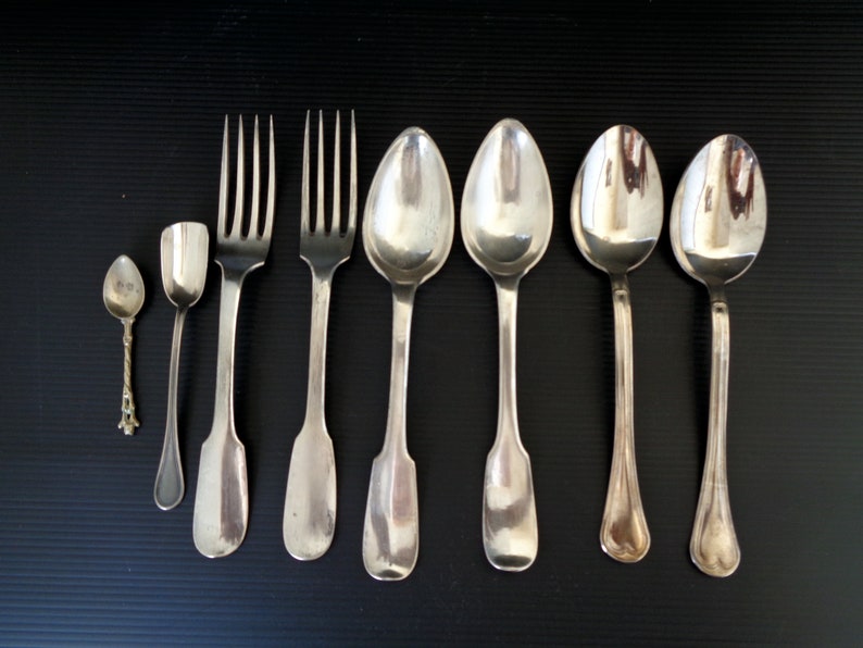May include: A set of seven silver-plated utensils, including two forks, four spoons, and a small spoon. The utensils are arranged in a row on a black surface.