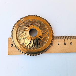 May include: A circular brass decorative plate with a scalloped edge and a central hole. The surface features a raised floral petal design. A wooden ruler is positioned underneath, showing the object has a diameter of approximately 3.5 inches.