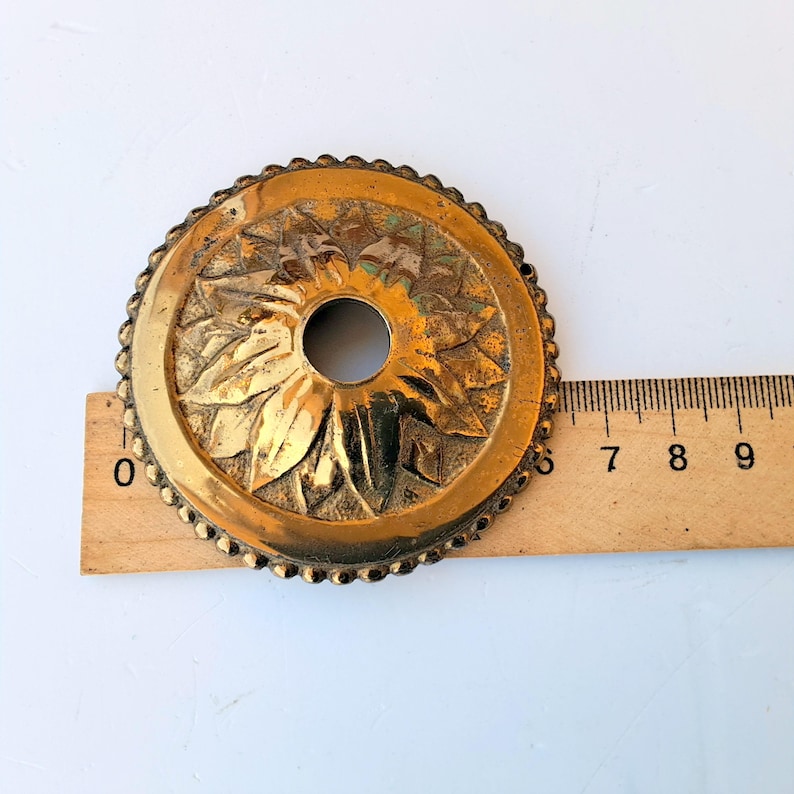 May include: A circular brass decorative plate with a scalloped edge and a central hole. The surface features a raised floral petal design. A wooden ruler is positioned underneath, showing the object has a diameter of approximately 3.5 inches.