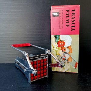 Pode incluir: Um cortador de batata vintage com uma alça vermelha e uma grade vermelha. O cortador de metal está ao lado de uma caixa com as palavras "TRANCIA PATATE" em vermelho. A caixa tem uma ilustração de vegetais.