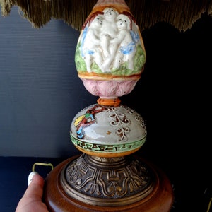 Op de afbeelding: Een vintage keramische tafellamp met een roze en witte basis versierd met cherubijnen. De lamp heeft een groene en gouden bol en een houten basis met een gouden metalen rand.