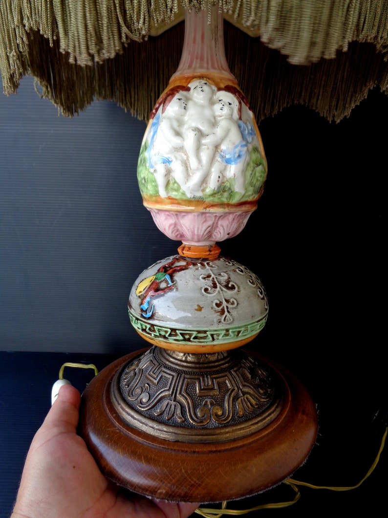 Op de afbeelding: Een vintage keramische tafellamp met een roze en witte basis versierd met cherubijnen. De lamp heeft een groene en gouden bol en een houten basis met een gouden metalen rand.