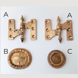 May include: Two antique brass hinges and two brass drawer pulls. The hinges are rectangular with a decorative center. One drawer pull is round with a central design and a laurel wreath border. The other drawer pull has a handle.
