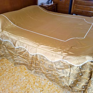 May include: A gold-coloured bedspread with white trim and a patterned skirt. The bedspread is draped over a bed, with a wooden headboard and furniture visible in the background. The bedspread has a satin-like sheen.
