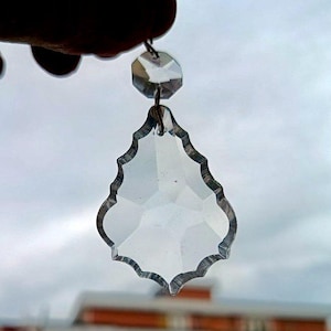 Könnte beinhalten: Ein klarer, facettierter Kristall-Kronleuchter-Anhänger mit dekorativem, gewelltem Rand. Der Anhänger ist an einem kleinen, achteckigen Kristall aufgehängt. Der Hintergrund ist eine verschwommene Ansicht eines bewölkten Himmels und eines Gebäudes.