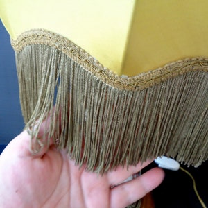 Op de afbeelding: Een gele lampenkap met een gouden rand en franje. De franje is gemaakt van een dun, bruin materiaal.