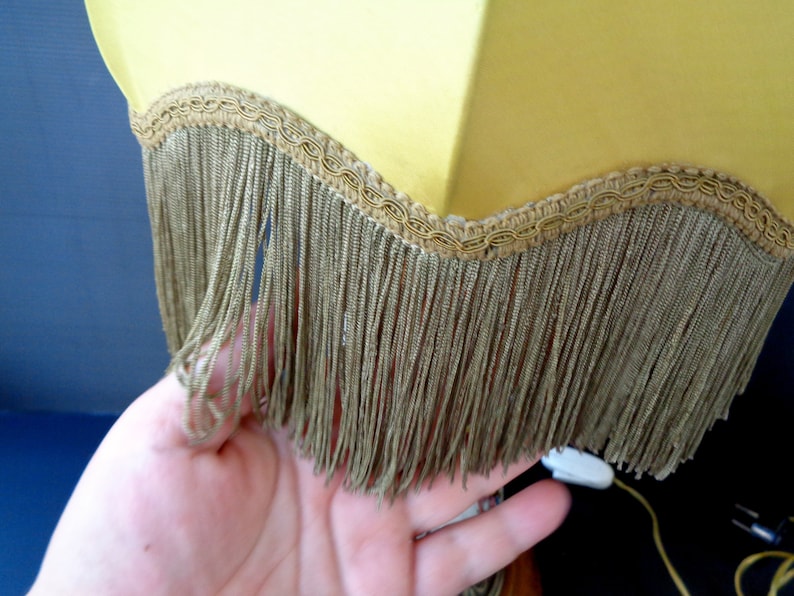 Op de afbeelding: Een gele lampenkap met een gouden rand en franje. De franje is gemaakt van een dun, bruin materiaal.