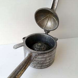 May include: A vintage, silver-colored citrus juicer with a hinged top and a ribbed base. The juicer has a handle for squeezing and a perforated reamer inside. The top is open, revealing the interior.