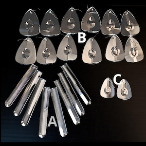 Op de afbeelding: Een verzameling heldere glazen prisma hangers en staven. De hangers zijn driehoekig met een cirkelvormig ontwerp in het midden. De staven zijn rechthoekig met afgeronde uiteinden, op een zwarte achtergrond.