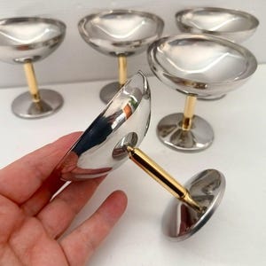 Peut inclure: Ensemble de cinq coupes à champagne. Les verres présentent une coupe argentée, une tige dorée et un pied argenté. L'un des verres est tenu, mettant en valeur le design. Les verres sont sur une surface blanche.