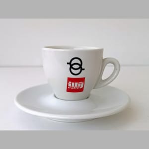 Può includere: Set tazza e piattino da espresso bianchi. La tazza presenta un logo nero e rosso. Il piattino è rotondo e ha un centro incassato per tenere la tazza. Il set è in ceramica.