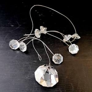 May include: Clear crystal chandelier prisms of various sizes are strung on thin, clear cords. The largest prism is a faceted, round shape. The smaller prisms are octagonal and irregular shapes. The prisms are displayed against a dark background.