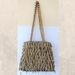 Puede incluir: Bolso de mano beige con cuentas y asas a juego. El cuerpo del bolso es de malla, adornado con cuentas de madera verticales. Las asas están hechas de pequeñas cuentas redondas. El bolso tiene forma rectangular.