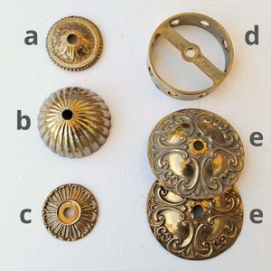 May include: A collection of brass hardware components labeled a through e. Item a is a small floral canopy, b is a fluted dome, c is a flat circular plate, d is a cylindrical ring with side holes, and e shows two ornate circular plates with scrollwork.