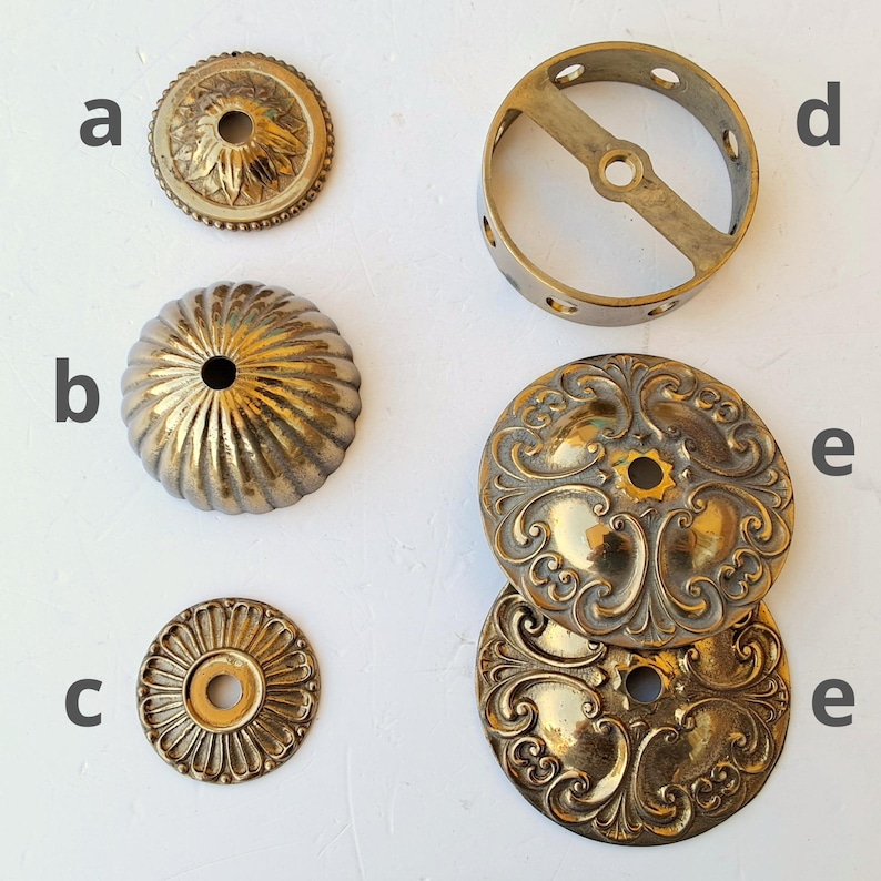 May include: A collection of brass hardware components labeled a through e. Item a is a small floral canopy, b is a fluted dome, c is a flat circular plate, d is a cylindrical ring with side holes, and e shows two ornate circular plates with scrollwork.