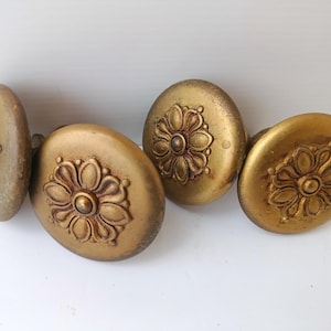 May include: A collection of five antique gold-colored cabinet knobs. Each oval knob features a raised floral design in the center. The knobs have a distressed finish, suggesting age and use. These decorative knobs are suitable for furniture restoration.