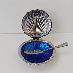 Puede incluir: Una caja de baratijas plateada en forma de concha con tapa abisagrada, que revela un interior azul vibrante. Una pequeña cuchara plateada descansa dentro de la caja. La caja tiene un borde festoneado.