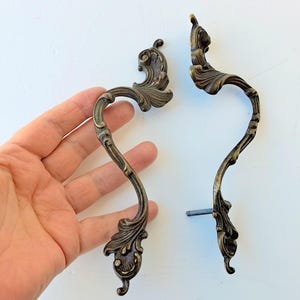 May include: Two antique bronze-colored cabinet handles with ornate, curved designs. Each handle features a scrolling leaf pattern and a decorative finial. The handles are shown against a white background.